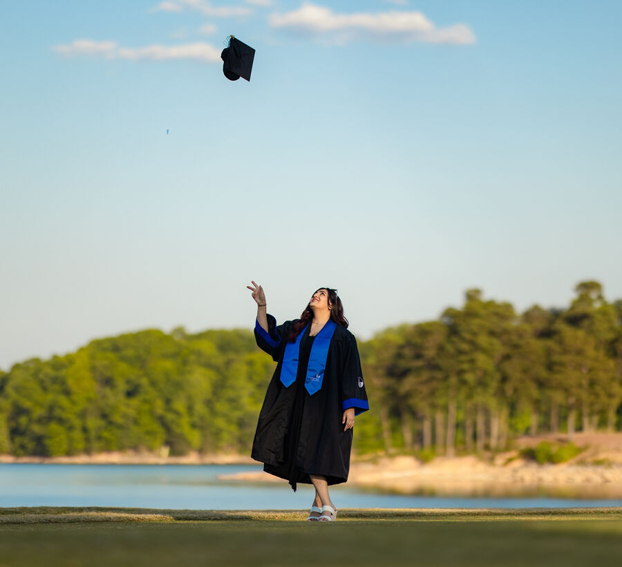Graduate in gown tossing her cap into the sky by a lake